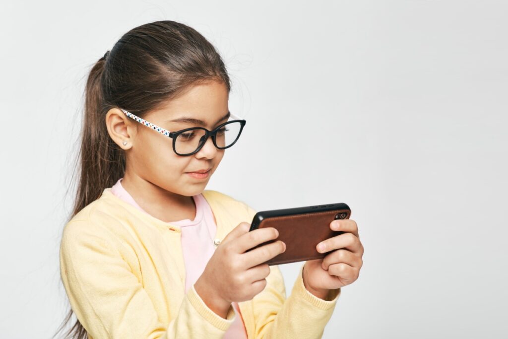 Young girl wearing black eyeglasses concentrating while holding and using smartphone device in yellow cardigan.