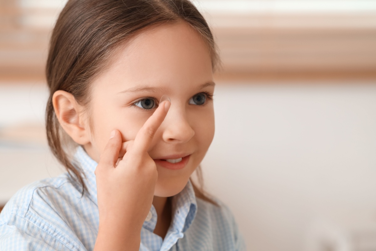 Young girl with long brown hair rubbing her eye showing signs of eye strain or vision discomfort in light blue shirt.